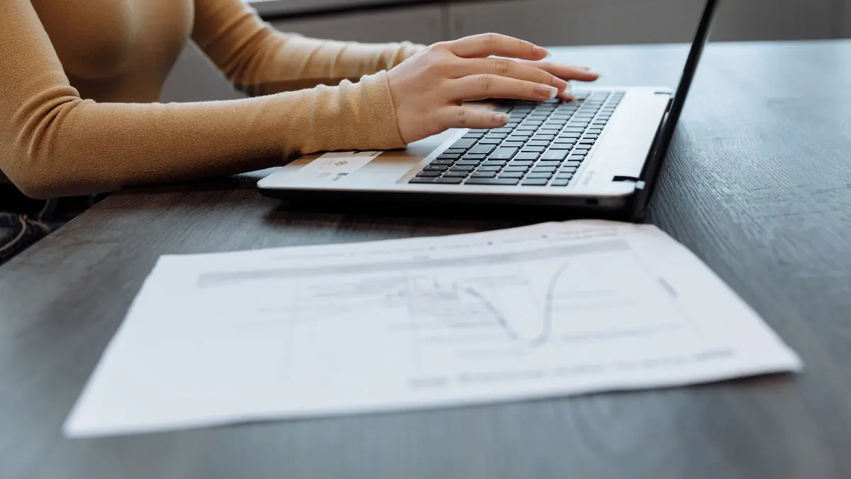 Close-up of a person's hands typing on a laptop at a desk with documents. Ideal for office, work, and technology themes.