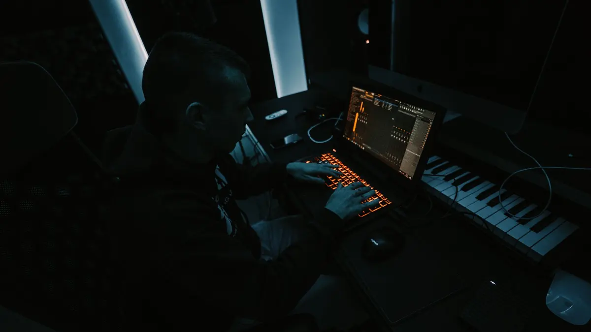 Music producer focused on composing tracks in a dimly lit studio with laptop and keyboard.